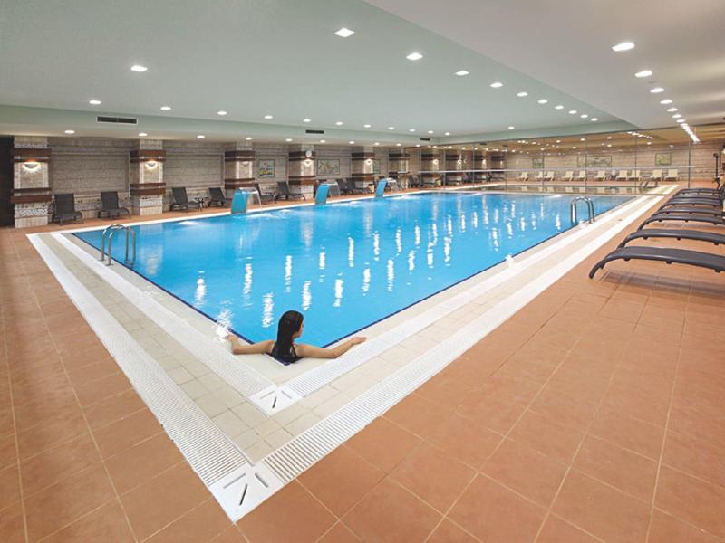 Großes Hallenbad mit klarem Wasser, weißem Rand und braunen Fliesen, Schwimmer im Becken.