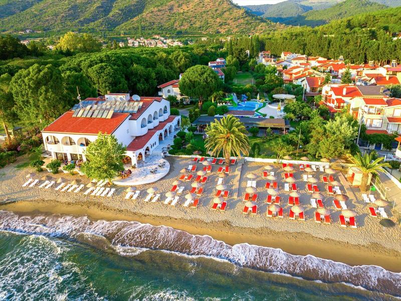 Beach hotel with red umbrellas and sun loungers in front of green hills and forested mountains.