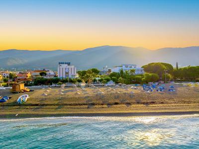 Strand mit blauem Wasser, Sand, Sonnenschirmen und Gebäuden vor Bergen im Sonnenuntergang.