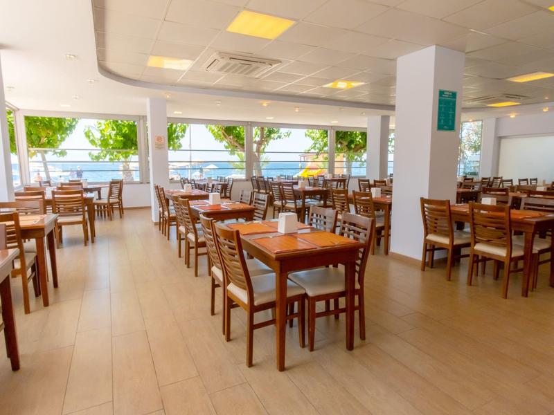 Světla a prostorná restaurace s mnoha dřevěnými stoly a židlemi u velkých oken s výhledem na stromy.