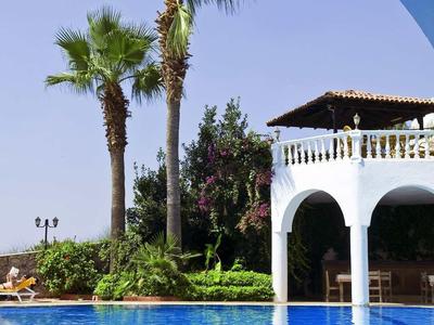 Blauer Pool vor weißem Gebäude mit Säulen und hohen Palmen unter klarem Himmel.