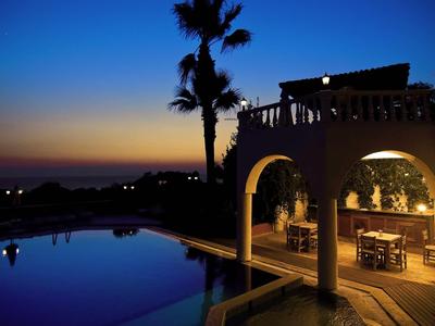 Abendliche Poollandschaft mit Palmen und beleuchtetem Haus unter dunklem Himmel.