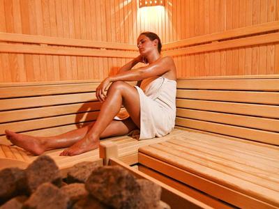 Frau in weißem Handtuch sitzt entspannt in hellem Holzsauna mit Steinen im Vordergrund.