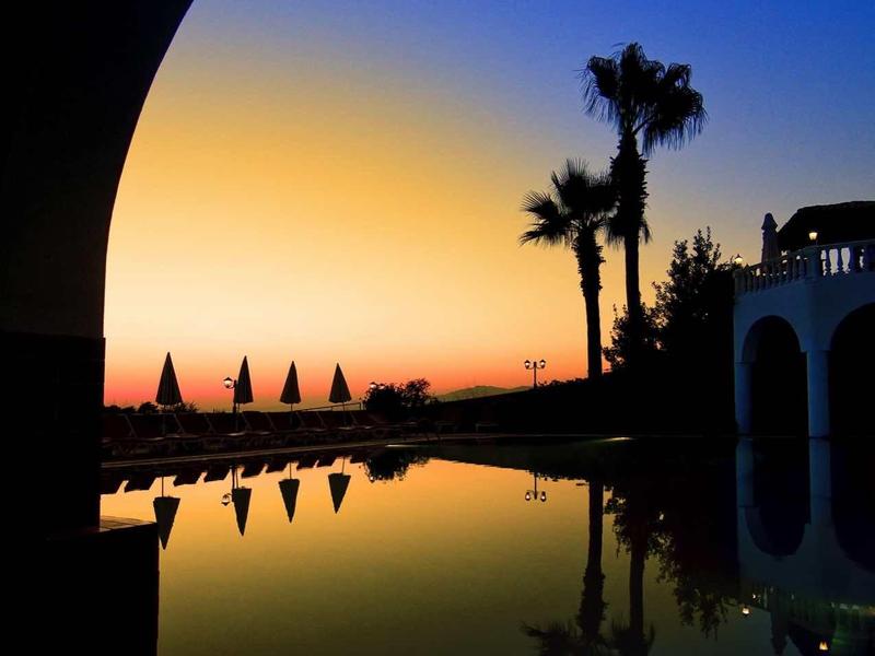 Sonnenuntergang mit Palmen und Sonnenschirmen, reflektiert in ruhigem Wasser, Blick durch einen Bogen.