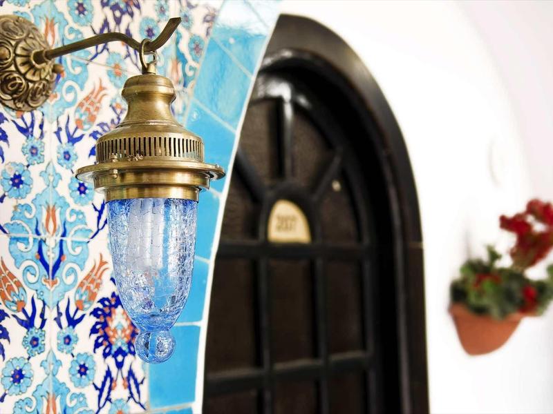 Wandlampe mit blauem Glas und Messing, neben bunter Fliesenwand und schwarzer Holztür mit Blumen.