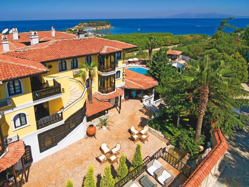 Großes gelbes Haus mit roten Dächern, Terrasse, Pool, Palmen und Blick aufs blaue Meer im Hintergrund.