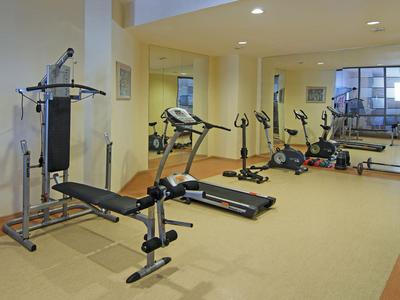 Salle de sport lumineuse avec bancs de musculation, tapis de course et vélo d'appartement devant un grand miroir.