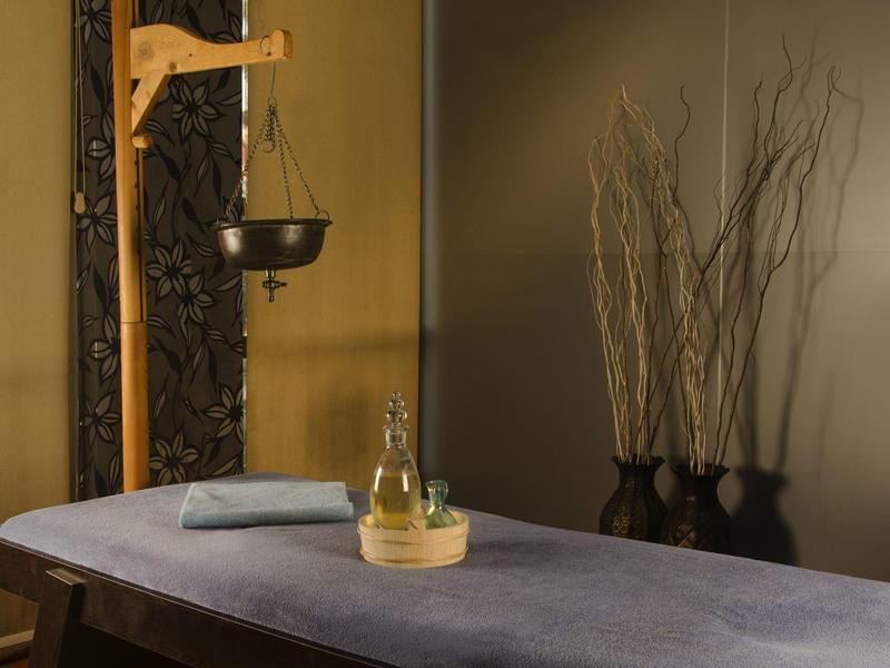 Uklidňující masážní místnost s lehátkem, oleji a dekorativními větvemi v pozadí.