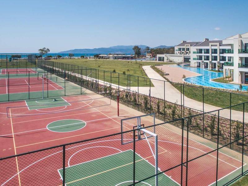 Meerdere tennisbanen naast een hotel met zwembaden en wandelpaden onder een heldere lucht.