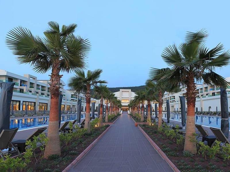 Hotelresort met twee zwembaden, palmbomen en een centrale wandelweg bij heldere lucht.