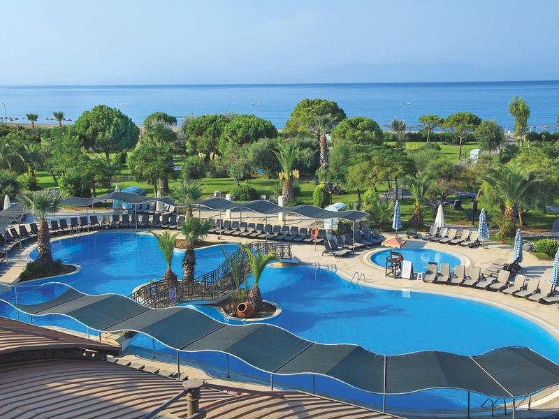 Large hotel pool with loungers and sea view, surrounded by palm trees and green landscape.