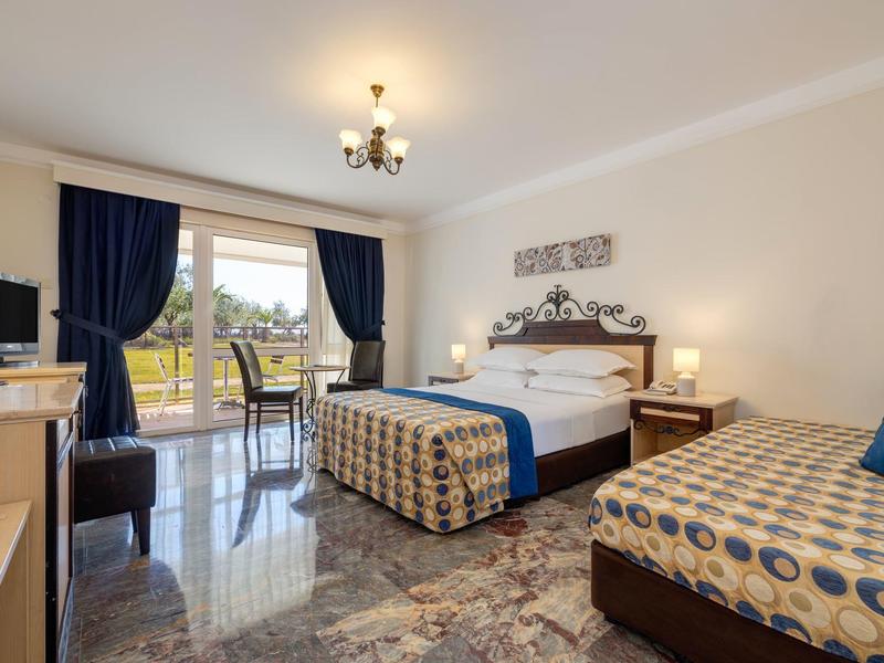 Bright hotel room with double bed, single bed, balcony, and blue-gold decor.