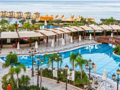Großer Hotelpool mit weißen Liegen, Palmen und Blick auf das Meer mit Strandbungalows.