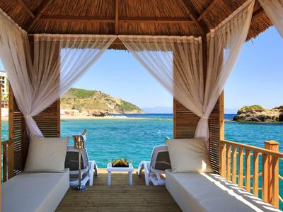 Offene Holzlaube mit weißen Vorhängen, zwei Liegen und Meerblick auf Felsen und Hügel.