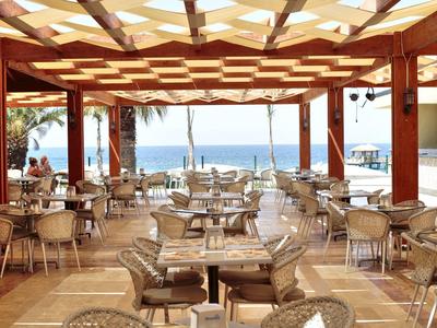 Offener Restaurantbereich mit Holzdecken, vielen Tischen und Blick aufs blaue Meer.