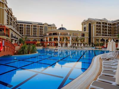 Großer Swimmingpool mit blauen Fliesen vor einem mehrstöckigen Hotelkomplex bei Sonnenuntergang.