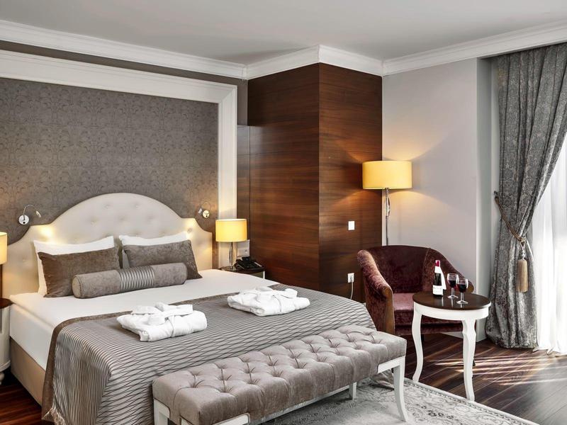 Schlafzimmer mit grauem Bett, gepolsterter Bank, Nachttischen, Sesseln und hellem Vorhang.