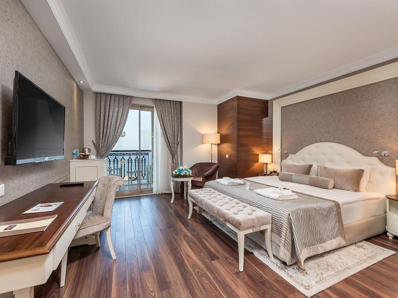 Elegantes Hotelzimmer mit Holzboden, Doppelbett, Schreibtisch, Fernseher und Balkon mit Vorhängen.