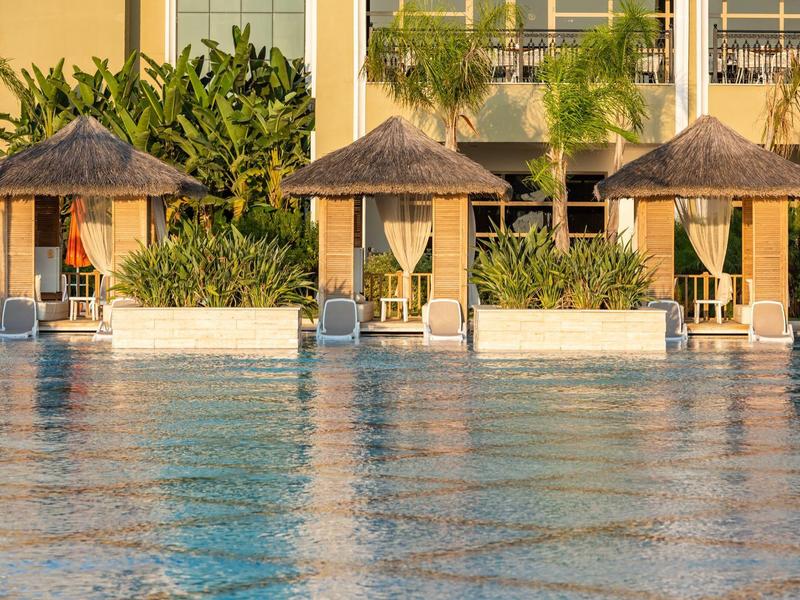 Pool mit drei Strohdach-Sonnenschirmen, Liegestühlen und tropischen Pflanzen vor gelbem Gebäude.