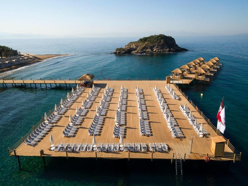 Große Holzplattform mit Liegestühlen und Sonnenschirmen über blauem Meer und kleinem Inselchen.