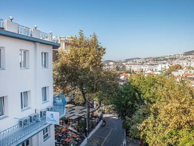 Blick auf eine Straße mit Bäumen, weißen Gebäuden und einer Terrasse bei klarem Himmel.