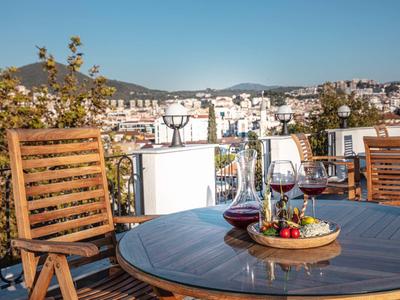 Terrasse mit Holzmöbeln, Weingläsern und Obstschale vor Stadt- und Bergkulisse bei klarem Himmel.