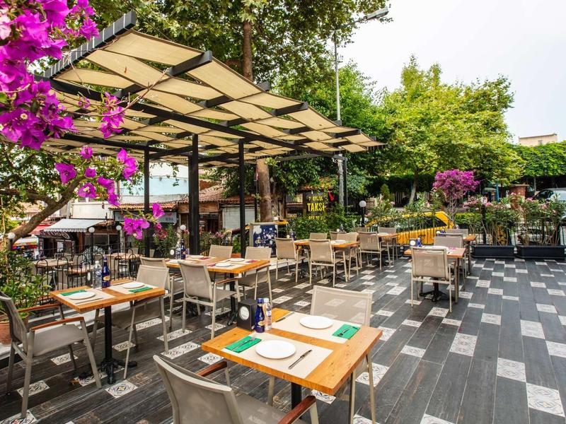 Große Terrasse eines Cafés mit Holztischen, Stühlen, blühenden Pflanzen und Sonnensegeln.