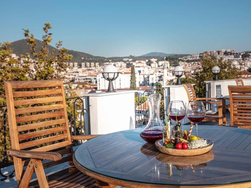 Terrasse mit Holzmöbeln, Weingläsern und Obstschale vor Stadt- und Bergkulisse bei klarem Himmel.