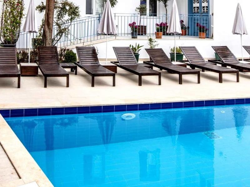 Reihe brauner Liegestühle neben klarem blauen Pool, weiße Terrasse und Pflanzen im Hintergrund.