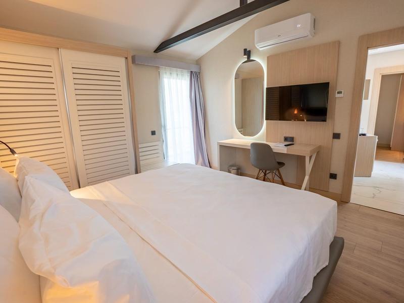 Helles Schlafzimmer mit Doppelbett, Schrank mit weißen Türen, Schreibtisch, Spiegel und TV an der Wand.