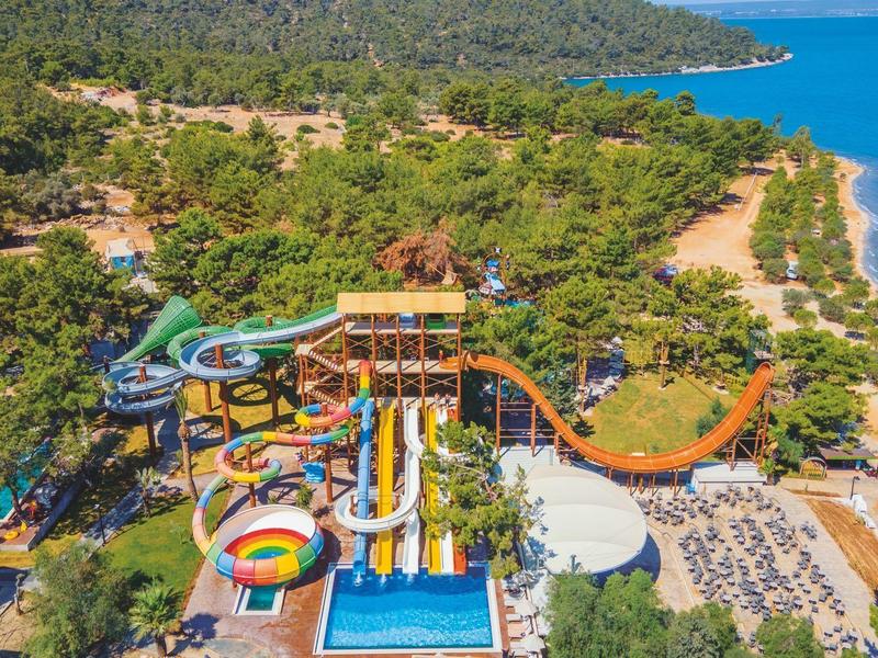 Wasserpark mit farbigen Rutschen, Pool und Strand in grüner, bewaldeter Küstenlandschaft.