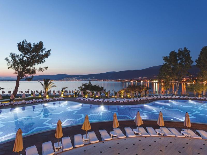 Großer Pool mit blauer Beleuchtung, umgeben von Liegestühlen und Sonnenschirmen am Meer bei Abenddämmerung.