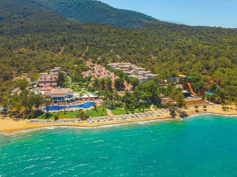 Küste mit türkisfarbenem Meer, Sandstrand, Hotels und Wald im Hintergrund, Berge am Horizont.