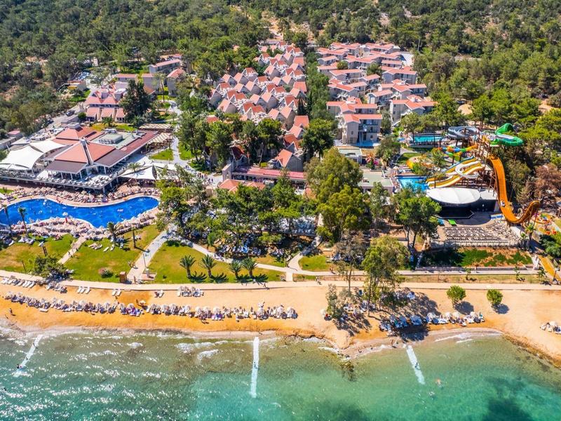 Luftaufnahme eines Resorts mit Pools, Wasserrutschen, Strand und klarem Meerwasser in grüner Umgebung.