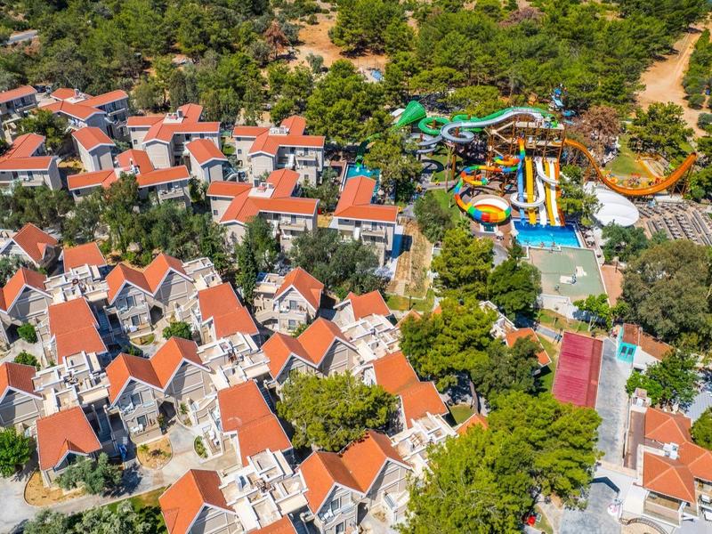 Luftaufnahme eines Ferienparks mit roten Dächern, Wasserpark, viel Grün und Strand am Meer.