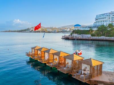 Kleine Holzhäuser am Wasser vor einem Hotel mit türkisem Meer und klarem Himmel.