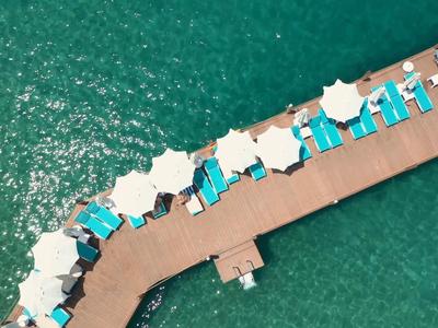 Luftaufnahme eines Piers mit weißen Sonnenschirmen und blauen Liegestühlen am türkisfarbenen Wasser.