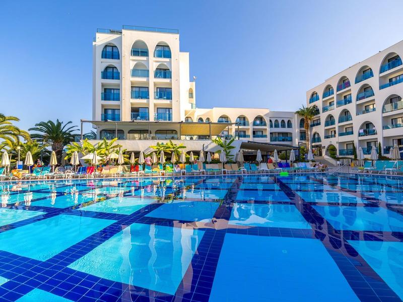 Großer Hotelpool mit klarem Wasser und umliegenden weißen Gebäuden unter blauem Himmel.