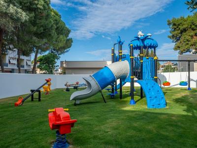Bunter Spielplatz mit Rutsche, Kletterwand, Wippe und Schaukeln auf grünem Rasen unter blauem Himmel.