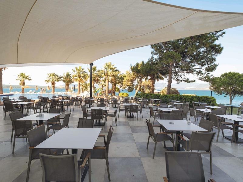 Venku posezení s stoly, výhled na moře a palmy pod bílým stanem.