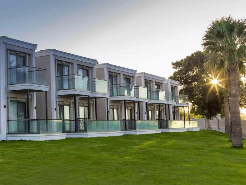 Moderní hotelová budova se skleněnými balkóny a zeleným trávníkem při východu slunce.