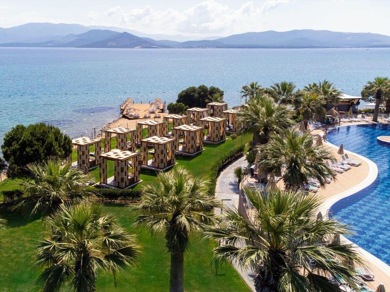 Rezort u moře s kabinami, palmami a velkým zakřiveným bazénem, v dálce hory.