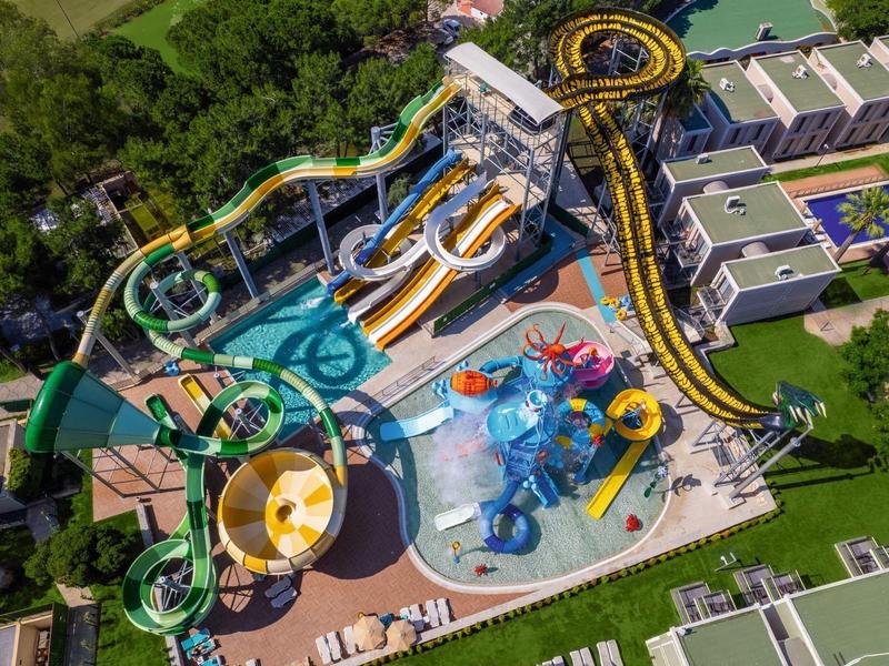 Letecký pohled na barevný vodní park s tobogány a bazény obklopený zelení a budovami.