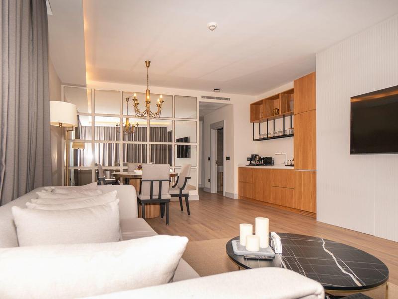 Moderní obývací prostor s béžovými pohovkami, kulatým černým konferenčním stolkem, dřevěnými skříňkami a jídelním stolem v pozadí.