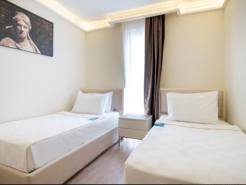 Jednoduchý hotelový pokoj se dvěma jednolůžky, bílým povlečením, oknem a minimalistickým vybavením.