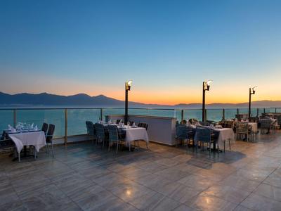 Freiluft-Restaurant mit gedeckten Tischen, Meerblick und Bergen bei Sonnenuntergang auf Terrasse.