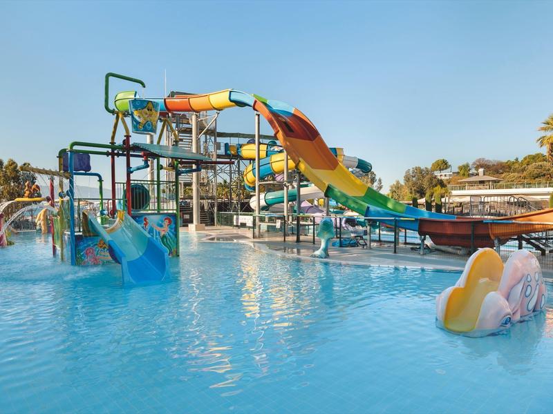 Colorful water slides and play structures in a large pool with mist and palm trees nearby.