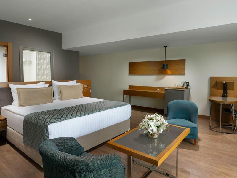 Spacious hotel room with double bed, sitting area, desk, and modern wooden furniture.