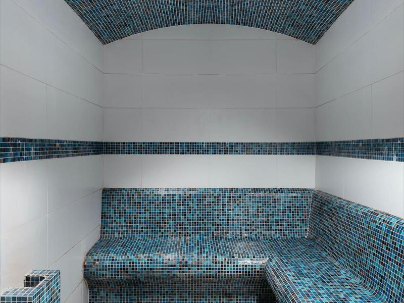 Small steam room with blue mosaic tiles and white floor tiles, bench along the walls.