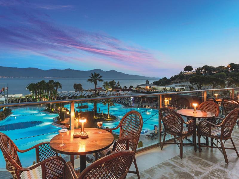Poolside dining area with tables, chairs, and lit candles overlooking a large pool and ocean at sunset.